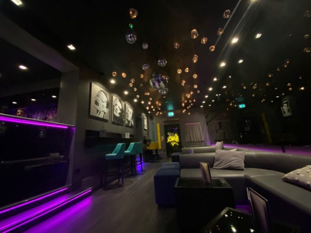 Interior of The Light Lounge gay bar. Stylish lighting, modern decor, bar area with purple accents, and comfortable lounge seating.