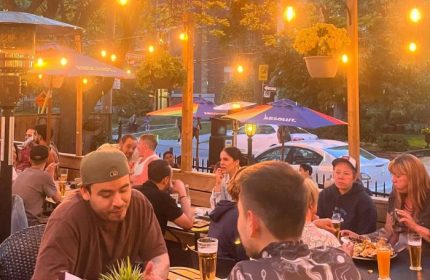 The Lodge at O'Grady's in Toronto: Outdoor patio scene with gay men enjoying drinks and food under string lights and rainbow umbrellas.