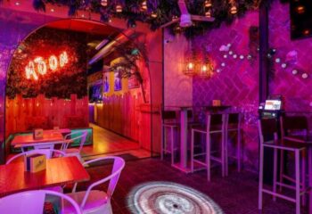 Interior of The Moon Tropical Gay Bar in Barcelona. Neon sign, plants, tables, chairs, and pink lighting create a vibrant atmosphere.