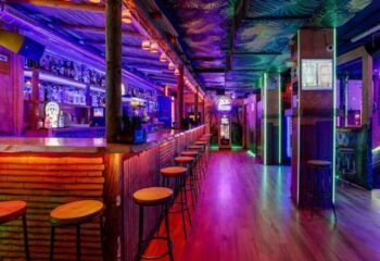 Interior of The Moon Tropical Gay Bar in Barcelona. Bar stools line the counter. Colorful lights illuminate the space.