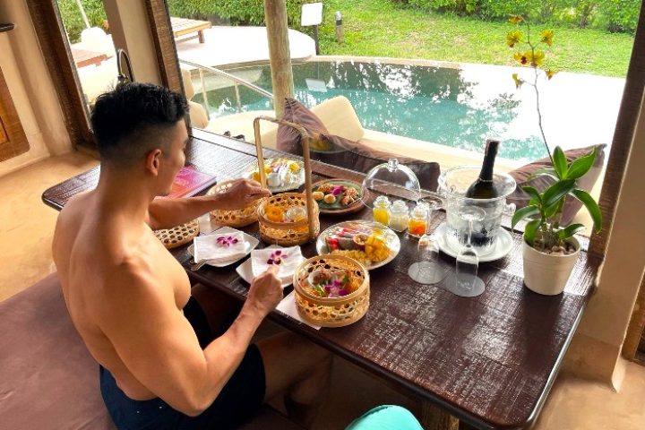 Gay man enjoys breakfast at The Naka Island, a Luxury Collection Resort & Spa, Phuket. Beachfront pool villa with wine and fruit.
