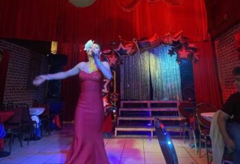 The New Jalisco Bar drag show: Drag queen performs in a red gown on stage with red curtains and star decorations. Gay travel advice.