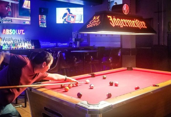 Man plays pool at The North End gay sports bar in Chicago. Jägermeister light fixture hangs above the red felt pool table. Gay travel.