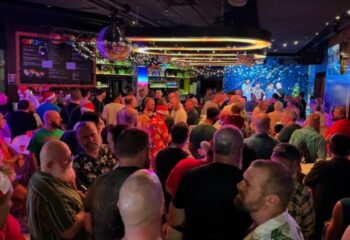 Crowded scene at The Oxford Hotel, Sydney. Gay bar with men, some wearing Santa hats, enjoying a festive event. Disco ball and stage visible.
