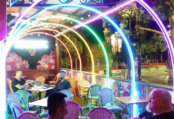 Outdoor seating at The Oxford Hotel Sydney, a gay-friendly travel destination. Rainbow lights arch over patrons. Nighttime scene.