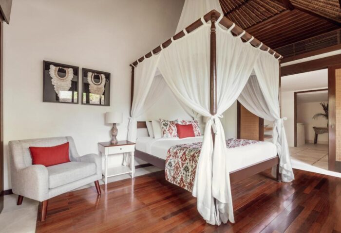 The Pavilions Bali hotel room. Gay-friendly hotel with a four-poster bed, white curtains, chair, and hardwood floors. Bali travel destination.