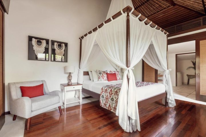 The Pavilions Bali hotel room. Gay-friendly hotel with a four-poster bed, white curtains, chair, and hardwood floors. Bali travel destination.