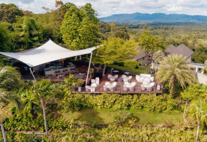 The Pavilions Phuket gay hotel view. Outdoor lounge area with white furniture, lush greenery, and mountain backdrop. Gay travel Phuket.