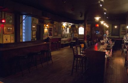 Interior of The Phoenix Gay Bar in New York. Bar stools line the counter. Walls are adorned with pictures. Dark lighting. Gay travel.