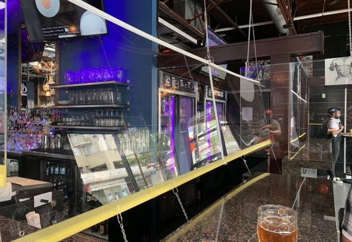 The Pumpjack Pub in Vancouver: A gay bar with safety dividers. Shelves of glasses, bar equipment, and a TV screen are visible. Travel advice.