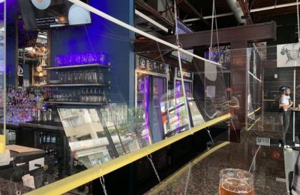 The Pumpjack Pub in Vancouver: A gay bar with safety dividers. Shelves of glasses, bar equipment, and a TV screen are visible. Travel advice.