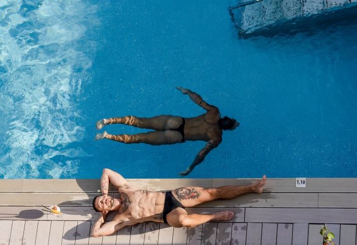 Two gay men at The Purple Hotel, Ibiza. One swims in the pool, the other sunbathes poolside. Gays-only travel destination.