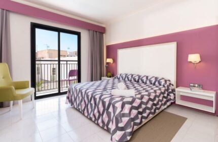 The Purple Hotel Ibiza room. Modern room in gay hotel, purple accent wall, balcony, geometric bedspread, and chair. Gay travel advice.