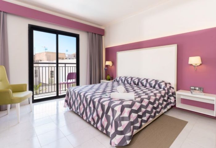 The Purple Hotel Ibiza room. Modern room in gay hotel, purple accent wall, balcony, geometric bedspread, and chair. Gay travel advice.