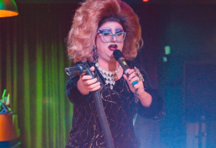 Drag queen performing at The Rem Bar, Manchester. Big hair, glasses, and jewelry. Holding a microphone and vacuum cleaner nozzle. Gay travel.