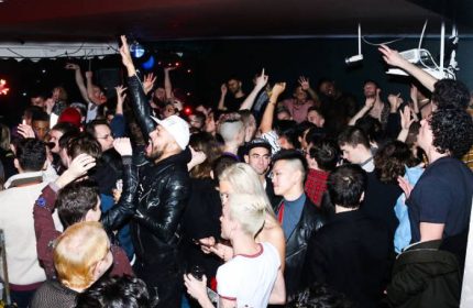 Packed crowd at The Rosemont, a chill gay bar in New York City. People dancing, raising hands, enjoying the nightlife scene.