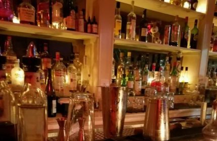 The Rosemont Chill, a gay bar in New York. Shelves of liquor bottles behind a bar with shakers and glassware. Gay travel advice.