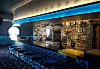 Interior of The Silver Fox Bar. Bar stools line a zigzag-patterned bar, with shelves of liquor bottles behind. Blue lighting adds a modern touch.