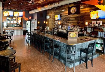 Interior of The SoFo Tap, a gay bear bar in Chicago. Long bar with stools, tables, TVs, and a rustic wood wall. Gay travel advice.