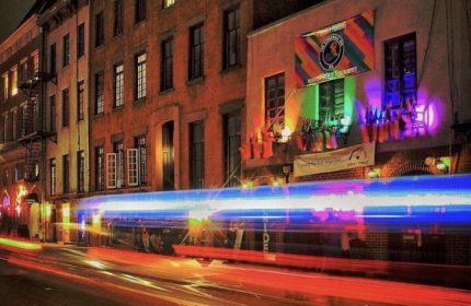 Night view of The Stonewall Inn in New York. Historic gay bar with rainbow flag, lights, and traffic streaks. Gay travel destination.