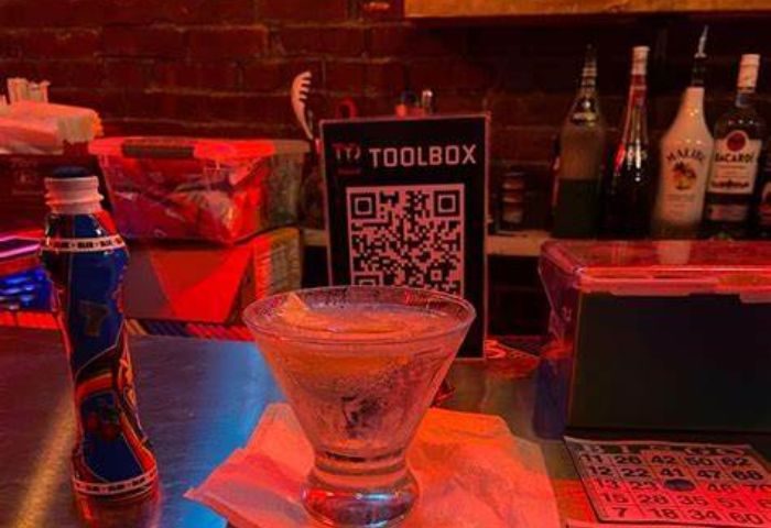 Toolbox Bar in New York: A cocktail on the bar with a Toolbox sign and QR code. Bottles, bingo card, and storage containers are visible. Gay bar.