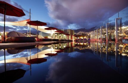 The Vine Hotel, a gay-friendly hotel in Madeira. Rooftop pool view at dusk, with city lights reflecting in the water. Loungers and umbrellas.