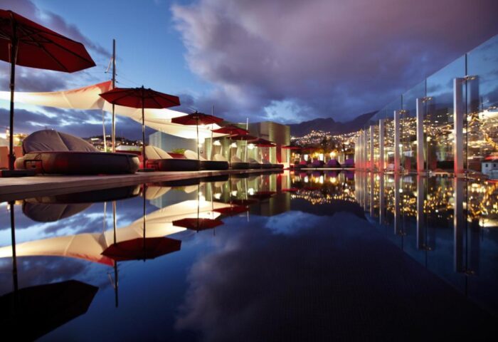 The Vine Hotel, a gay-friendly hotel in Madeira. Rooftop pool view at dusk, with city lights reflecting in the water. Loungers and umbrellas.