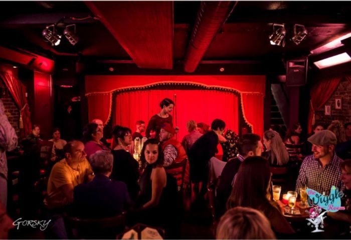 The Wiggle Room gay bar live show in Montreal. Red-lit interior with a stage, red curtains, and an audience enjoying the performance.