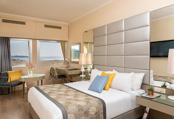 Leonardo Art Tel Aviv By the Beach hotel room. Modern decor, sea view, desk, and bed with blue and yellow pillows. Honeymoon hotel idea.