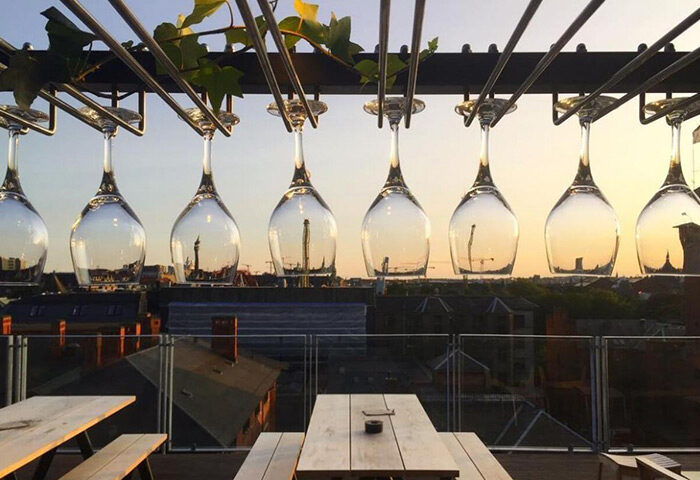 Hotel Danmark rooftop bar in Copenhagen's gayborhood. Wine glasses hang above tables, city skyline visible. A gay hotel in Vesterbro.