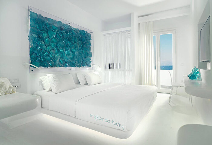 Mykonos Bay Resort & Villas beachfront hotel room. White interior, bed with Mykonos Bay logo, blue glass art. Gay honeymoon destination.