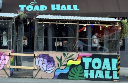 Toad Hall Bar in San Francisco. Gay bar exterior with rainbow art, flowers, and a man inside. Gay travel destination in San Francisco.
