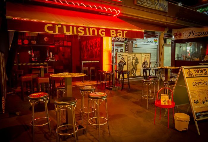 Tom's Cruising Bar, a gay cruise club in Gran Canaria. Exterior view with red lighting, bar stools, and COVID-19 protocol sign. Gay travel.