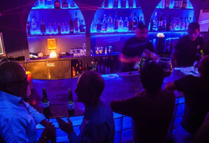 Tom's Leather Bar: Gay party in Mexico. Men drinking at the bar with blue lighting. Bartenders working, bottles on shelves.