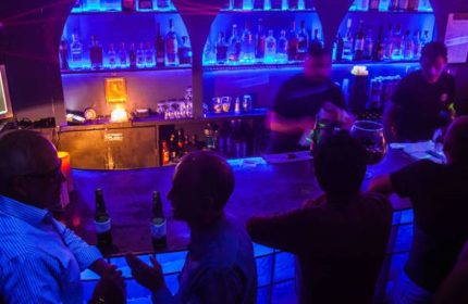 Tom's Leather Bar: Gay party in Mexico. Men drinking at the bar with blue lighting. Bartenders working, bottles on shelves.