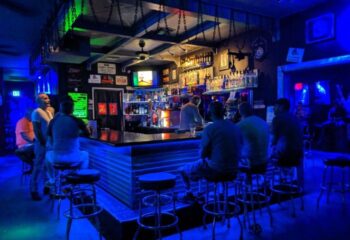 Interior of Tool Shed Bar, a gay bar. Men sit at the bar under blue lighting. Bottles and decor line the back. Gay travel advice.