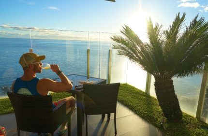 Gay man enjoying wine at Saccharum Hotel, Madeira. Ocean view, palm tree, sunny day. Top gay beach hotel on Instagram and TikTok. Portugal travel.