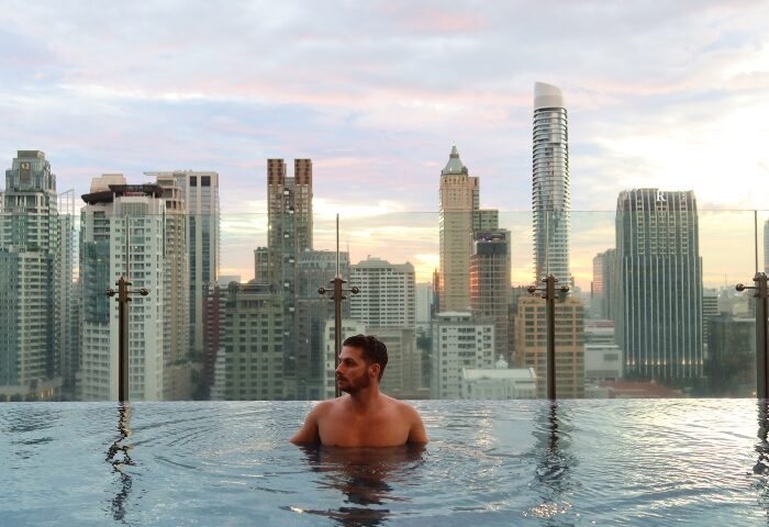 Gay honeymoon: Man in rooftop pool at Hotel Indigo Bangkok Wireless Road. Bangkok skyline view. Gay travel luxury.
