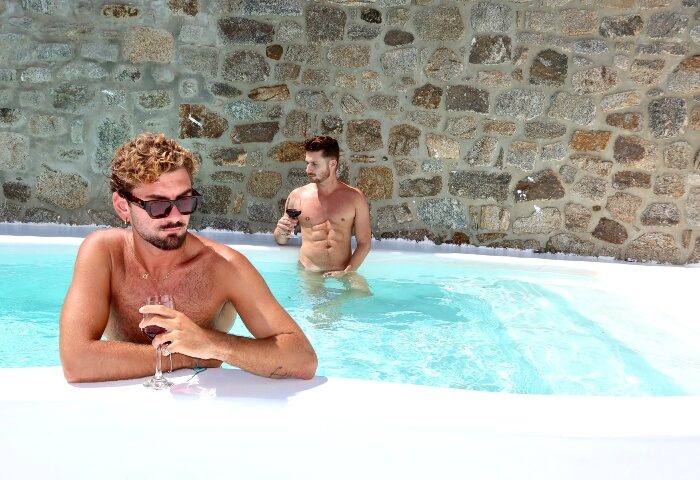 Gay honeymoon at Akkadian Gay Only Hotel Mykonos. Two men relax in a pool with wine, stone wall backdrop. Top gay pool villas.