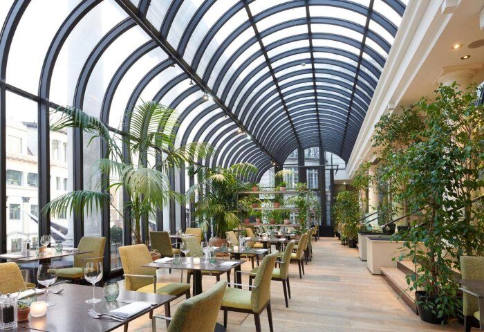 The Dilly's stunning dining area, a top gay honeymoon hotel in London's Soho. Tables are set beneath a soaring glass ceiling and lush greenery.