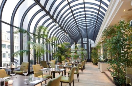 The Dilly's stunning dining area, a top gay honeymoon hotel in London's Soho. Tables are set beneath a soaring glass ceiling and lush greenery.