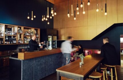 Saint Kilda Beach Hotel's bar in Melbourne. Bartender pouring drinks, patrons at tables. Top gay hotels with beach bar. Stylish lighting.