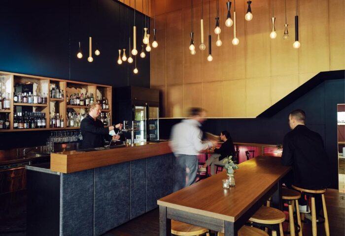 Saint Kilda Beach Hotel's bar in Melbourne. Bartender pouring drinks, patrons at tables. Top gay hotels with beach bar. Stylish lighting.