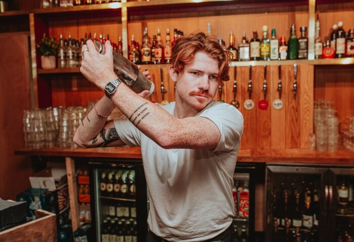 Gay bartender mixing drinks at Colours Hoxton, a top London gay dance club. Live DJ and vibrant nightlife for gay men's travel adventures.