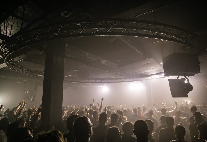 Crowd at BRÜT London, top monthly gay dance club. Hot men partying with hands in the air under lights and industrial club structure.