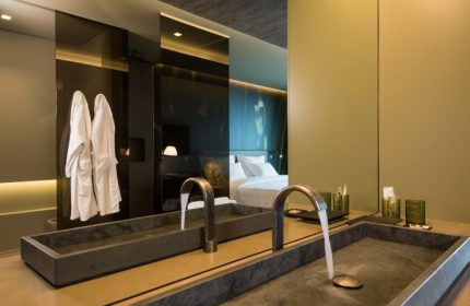 Saccharum Hotel Madeira bathroom with modern sinks, robes, and a mirror reflecting the bed. Top honeymoon beach hotel for mister and mister travel.