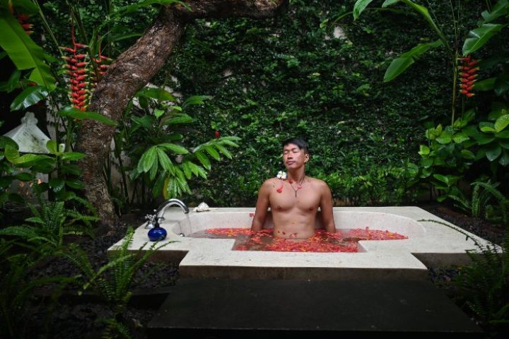 Gay wellness at The Pavilions Bali beach resort. Man relaxes in a flower-filled tub surrounded by lush greenery. Top gay travel destination.