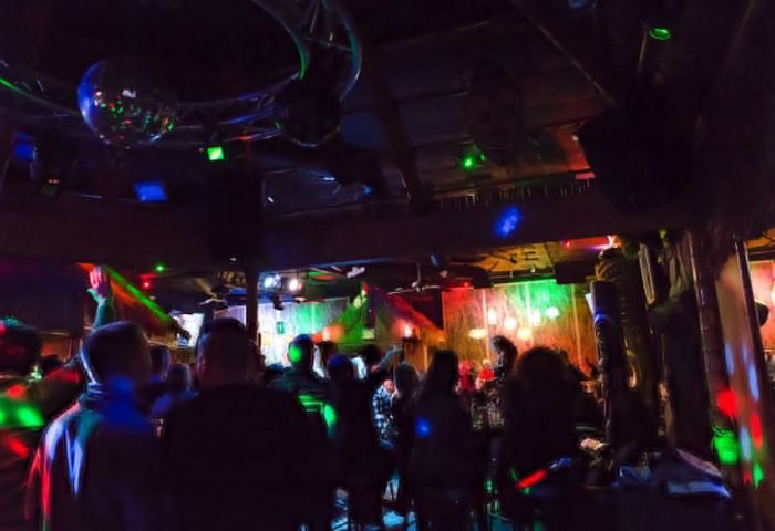 Crowded party at Toucans Tiki Lounge, a gay bar. Disco balls, colorful lights, and people mingling. Nightlife scene, gay travel destination.