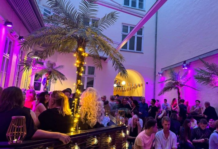 Trixie Copenhagen: A vibrant, crowded outdoor bar scene in Copenhagen, Denmark. Palm trees and purple lighting create a tropical vibe. Gay travel.