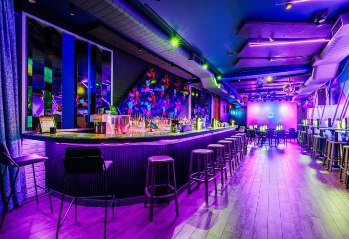 Interior of VERS, a gay venue in New York. Bar with stools, dance floor, stage, and colorful lighting. Nightlife travel advice.
